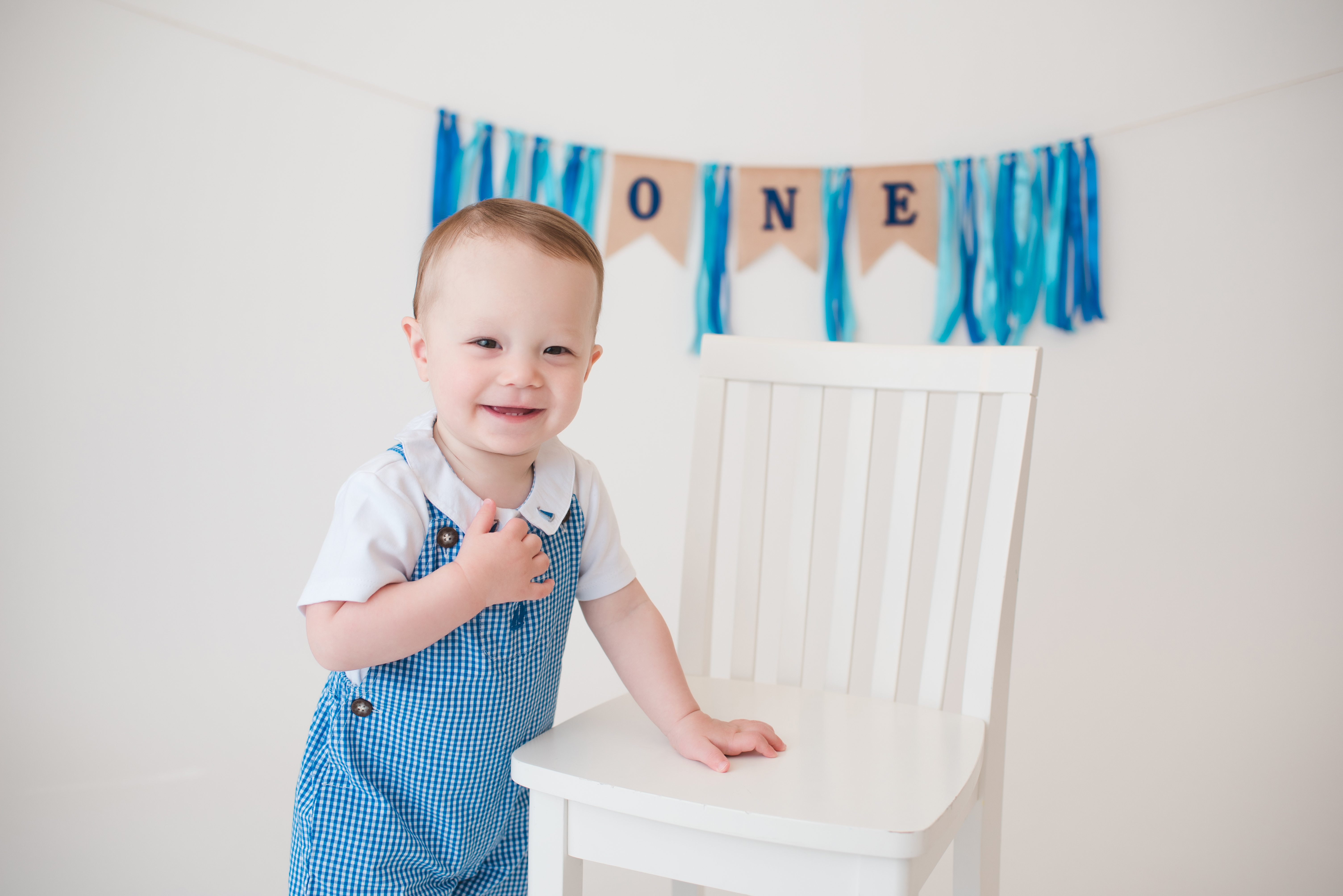 one year old wearing gingham romper, white child size chair, blue tassel banner