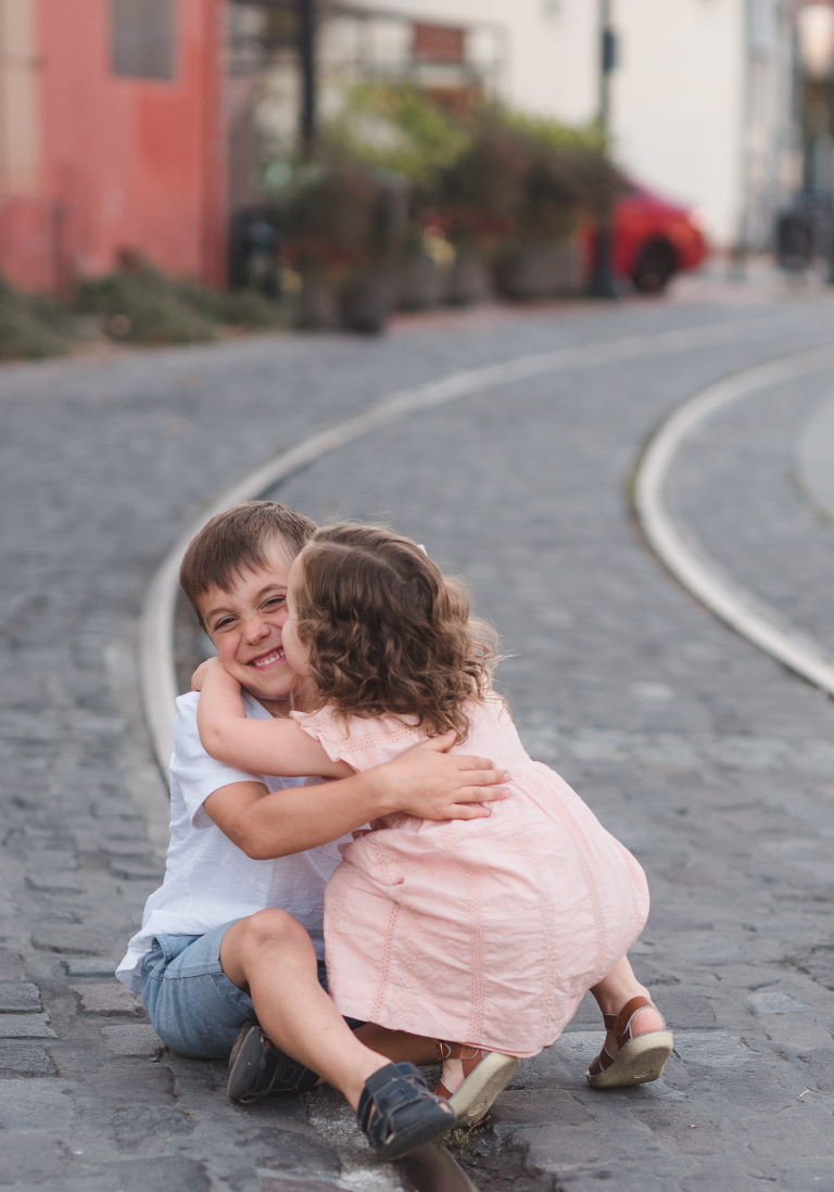 little girl kissing big brother on cheek, embracing siblings, hugging siblings, Keen sandals, Saltwater sandals, curly haired little girl, Petaluma cable car tracks, downtown Petaluma